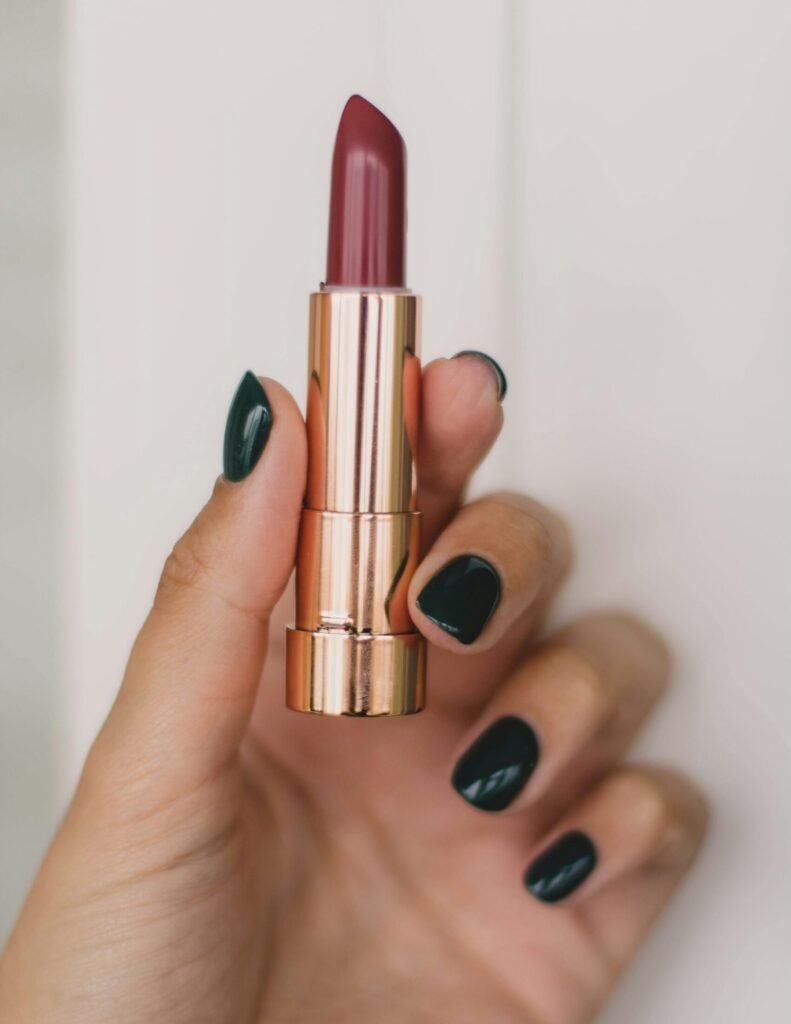 Close-up of a hand with dark polished nails holding a stylish lipstick tube against a neutral background.